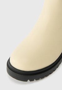 Béžová gumová bota s kulatou špičkou, hladkým povrchem a černou robustní podrážkou s výrazným vzorem na profilu.