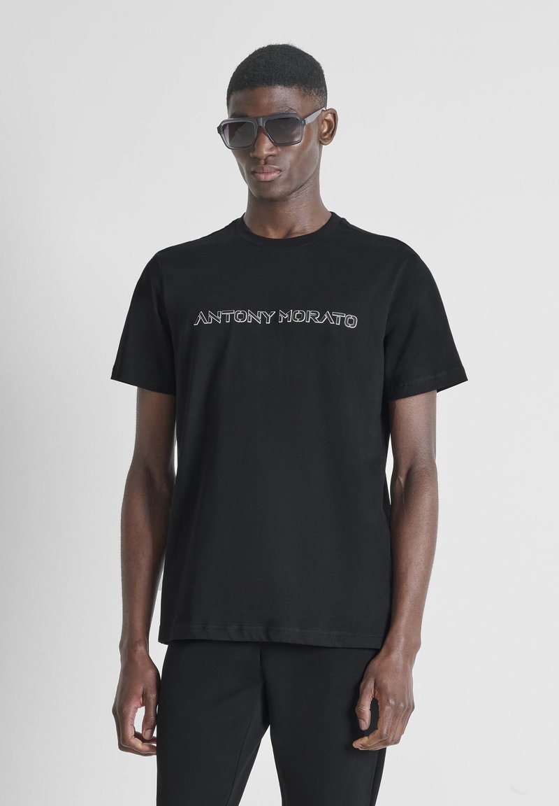 Mann trägt ein schwarzes Antony Morato T-Shirt und schwarze Hosen, posiert vor einem schlichten hellgrauen Hintergrund, accessoriert mit großen dunklen Sonnenbrillen.