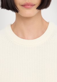 Maglione lavorato a maglia di colore crema con scollatura a coste, caratterizzato da un colore uniforme e una sottile trama sul tessuto. Vista ravvicinata.