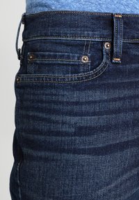 Tmavě modré denimové džíny s hladkým povrchem, standardním designem pěti kapes a viditelným šitím podél švů a pasu.