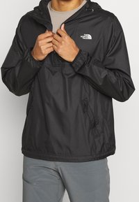 Persoon in een zwarte The North Face-jas met capuchon over een beige shirt, de rits aan het aanpassen, met grijze broek zichtbaar eronder.