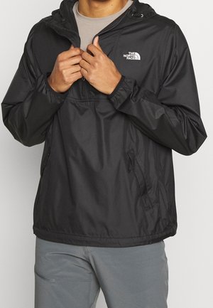 Persoon in een zwarte The North Face-jas met capuchon over een beige shirt, de rits aan het aanpassen, met grijze broek zichtbaar eronder.
