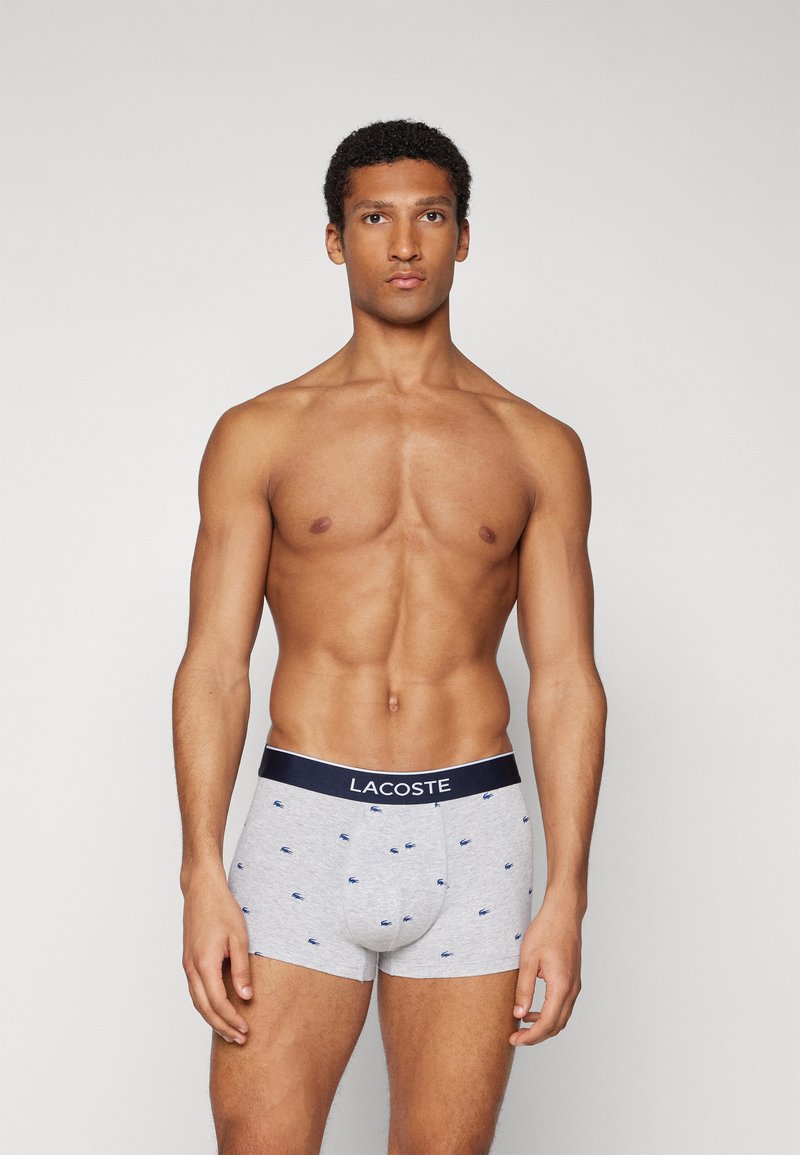 Szare bokserki z niebieskimi haftowanymi detalami, z granatową gumką z napisem "LACOSTE". Tkanina wygląda na miękką, z dopasowanym krojem.