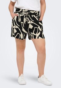 Persona que lleva pantalones cortos de patrón abstracto en negro y beige con cordón y zapatillas blancas, con las manos en los bolsillos.