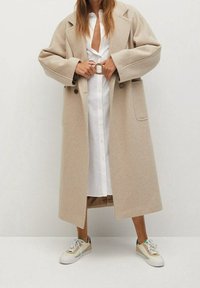 Beiger Wollmantel mit übergroßer Passform, großen Revers und Seitentaschen, getragen über einem weißen Hemdkleid, kombiniert mit mehrfarbigen Sneakers.