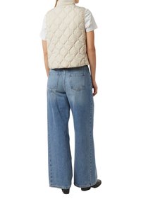 Béžová prošívaná vesta s vysokým límcem, nošená přes bílé tričko. Kombinováno s volnými světle modrými džínami a černou obuví.