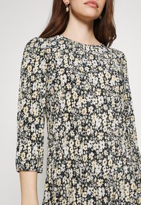 Blumendruckkleid mit schwarzem Hintergrund und gelben sowie weißen Blumen, dreiviertelärmeligen Ärmeln und strukturierter Stoff mit Raffungselementen.