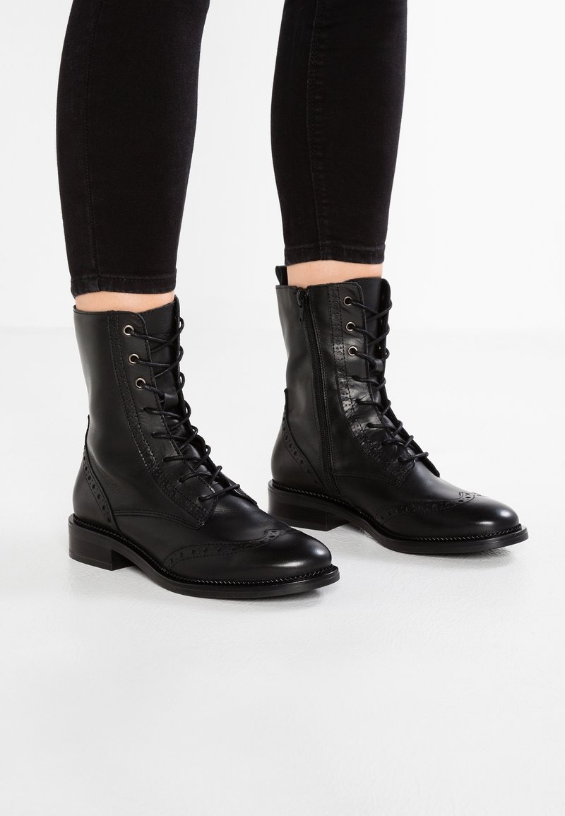Stivaletti in pelle nera alla caviglia con punta arrotondata, chiusura con lacci frontale, zip laterale, dettagli brogue e tacco basso. Superficie texturizzata e design elegante.