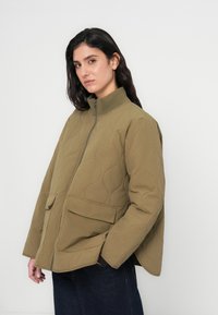 Πρασινοελιά, καπιτονέ μπουφάν με ψηλό φερμουάρ στον γιακά, δύο μπροστινές τσέπες και άνετη γραμμή, κατασκευασμένο από υφή, ανθεκτικό ύφασμα.