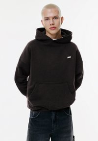 Dunkelbrauner Hoodie mit einer Vorderfernetasche, lockerer Passform und gerippten Ärmelbündchen. Beinhaltet ein kleines weißes Logo auf der Brust. Getragen mit dunklen Jeans.