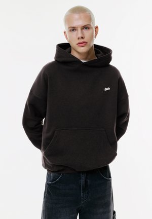 Haut à capuche marron foncé avec une poche avant, coupe ample et poignets côtelés. Comprend un petit logo blanc sur la poitrine. Porté avec un jean noir.