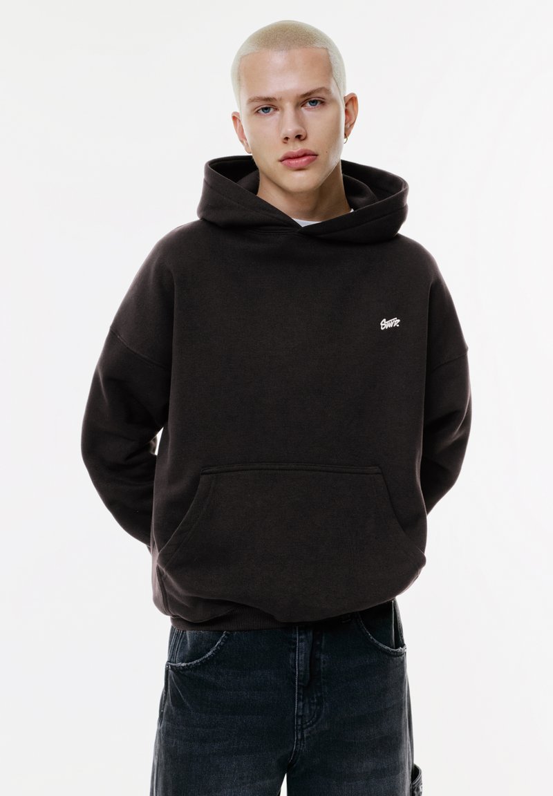 Dunkelbrauner Hoodie mit einer Vorderfernetasche, lockerer Passform und gerippten Ärmelbündchen. Beinhaltet ein kleines weißes Logo auf der Brust. Getragen mit dunklen Jeans.