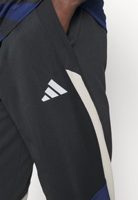 Schwarze Sporthosen mit beigen und marineblauen Akzenten. Verfügt über ein weißes Adidas-Logo auf dem linken Oberschenkel und besteht aus atmungsaktivem Material.