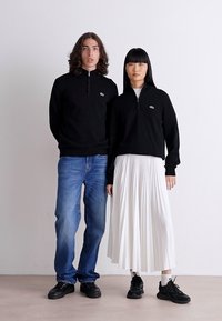 Pulover fleece negru cu fermoar pe stânga, asortat cu blugi; pe dreapta, un pulover scurt negru peste o fustă plisată albă, ambele purtând pantofi negri.