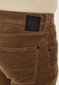 Brown corduroy pants featuring a textured fabric, slim fit, and a black label stitched on the back pocket with "RED POINT DENIM" branding.