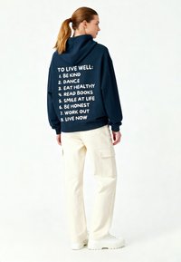 Hanorac bleumarin cu text alb pe spate, evidențiind opt principii de viață. Asortat cu pantaloni cargo crem și adidași albi.