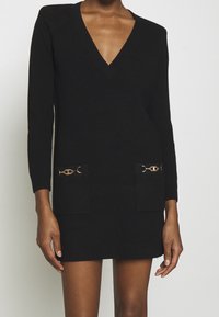 Abito maglione nero a coste con scollo a V, maniche lunghe, tasche frontali e accenti in hardware dorato. Tessuto morbido, design aderente.