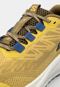 Yellow running shoe with a textured mesh upper, brown and blue lace accents, reflective details, and a white midsole designed for performance.