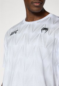 Άσπρο αθλητικό t-shirt με ανάγλυφο σχέδιο που απεικονίζει κύματα, μαύρο λογότυπο UFC στο αριστερό στήθος, στρογγυλή λαιμόκοψη και κοντά μανίκια.