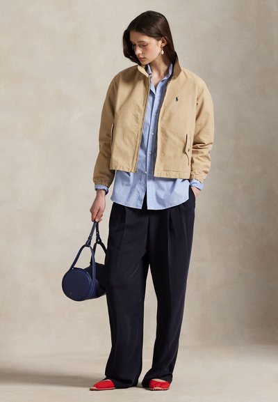 Kobieta w beżowej kurtce na niebieskiej koszuli, czarne szerokie spodnie, czerwone buty, trzyma granatową okrągłą torebkę, stoi na neutralnym tle.
