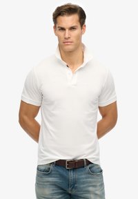 Weißes Poloshirt mit kurzen Ärmeln, einem Kragen, subtiler Textur und einem kontrastierenden Markendetail unter dem Kragen, kombiniert mit blauen Denim-Jeans.