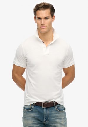 Polo shirt bianco a maniche corte con colletto, texture sottile e un dettaglio del marchio a contrasto sotto il colletto, abbinato a jeans in denim blu.
