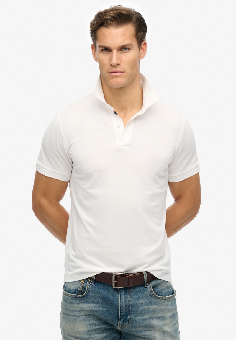 Weißes Poloshirt mit kurzen Ärmeln, einem Kragen, subtiler Textur und einem kontrastierenden Markendetail unter dem Kragen, kombiniert mit blauen Denim-Jeans.