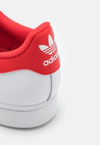 Zapatilla Adidas roja y blanca que presenta una parte superior de cuero sintético, collar de malla, cordones planos y una suela exterior de goma texturizada. Logo en el talón.