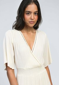 Robe cache-cœur couleur crème avec un texture plissée, manches courtes évasées, décolleté en V profond et couture décorative le long de l'encolure.