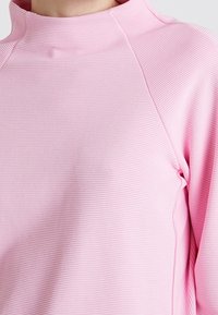 Růžový top s dlouhým rukávem a vysokým límcem, s texturovaným ribbovaným vzorem. Hladká látka se švy po stranách pro přizpůsobený střih.
