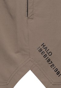 Beige shorts med en dragkedjficka, sömnadsdetaljer och texten: "HALO 19681971981" i svart på sidan. Slät tygtextur.