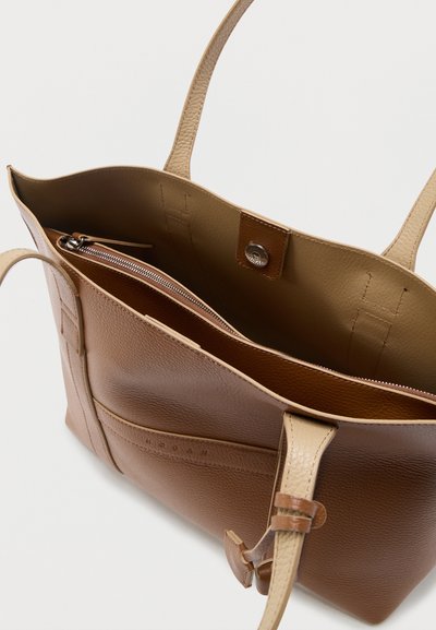 Sac fourre-tout en cuir marron et beige avec deux poignées, un compartiment principal ouvert et une poche intérieure zippée visible à l'intérieur.