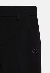 Juodos kelnės, pagamintos iš tekstūruoto audinio, su liesu siluetu. Yra priekinės kišenės ir diskretiškai išsiuvinėtas "CK" logotipas šone.