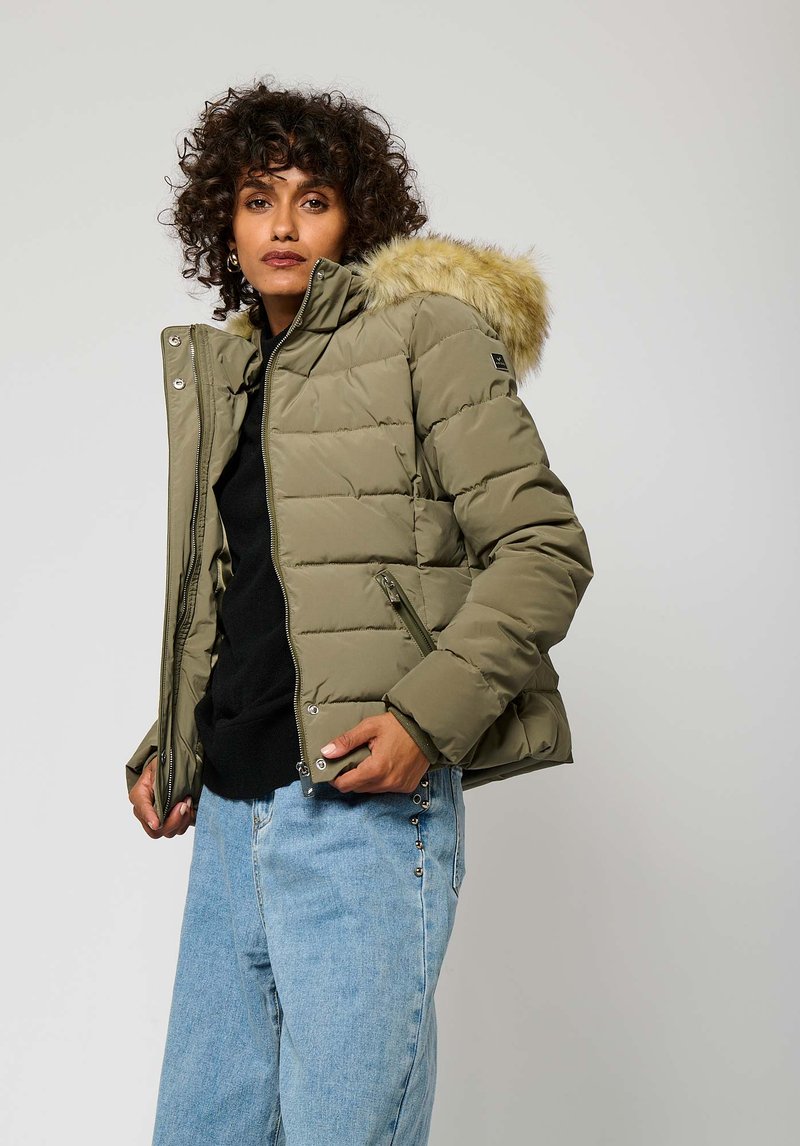 Olijfgroen pufferjack met een nepbontkap, ritssluiting en zijzakken, gedragen over een zwarte trui en lichtblauwe jeans.