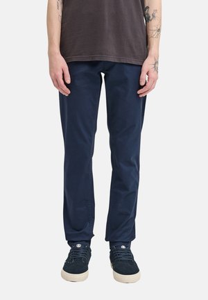 Osoba ubrana w granatowe spodnie, ciemne trampki z białymi podeszwami oraz ciemny krótki rękaw, stojąca z rękami swobodnie opuszczonymi wzdłuż ciała.