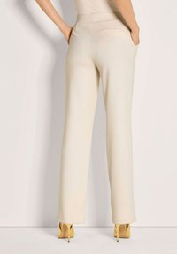 Vrouw in crèmekleurige rechte broek met handen in de zakken en gele hoge hakken, staande tegen een effen achtergrond.