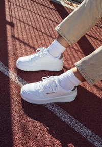 Sneaker bianco con una superficie in pelle liscia, suola testurizzata e dettagli in argento. Abbinato a calze bianche a coste e pantaloni beige con risvolto, su una pista rossa.