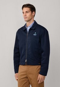 Giacca blu navy con chiusura a zip e colletto. Presenta un piccolo logo ricamato e tasche laterali. Abbinata a pantaloni khaki.
