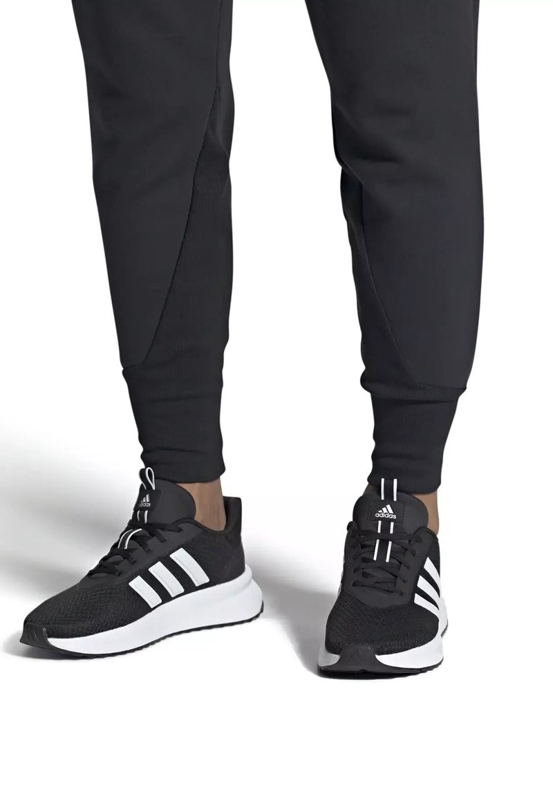 Personne portant des chaussures de course Adidas noires avec des bandes blanches et un pantalon de jogging noir, debout sur une surface blanche.