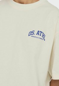Cremefarvet t-shirt lavet af bomuld, med rund halsudskæring og korte ærmer. Trykt blå tekst står der "OS. ATHL VERSATILE HYBRIDS."