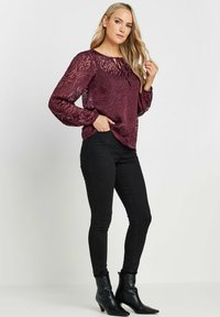 Blusa trasparente color cranberries con stampa animale e maniche a sbuffo, abbinata a jeans neri aderenti e stivaletti alla caviglia. Tessuto morbido, vestibilità rilassata.