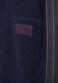 Casaco de fleece azul-marinho com superfície texturada, apresentando um rótulo roxo com "70" e um design listrado em roxo escuro com detalhes amarelos.