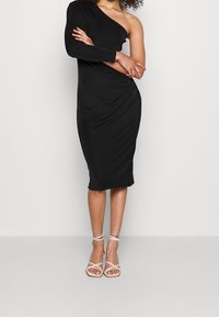 Nainen, jolla on ykkösvaihtoehto musta midi-mekko ja valkoiset remmikorkokengät, seisoo kädet ristissä.