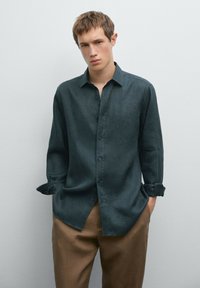 Dark green long-sleeve shirt made of a lightweight fabric, featuring a button-up front, a classic collar, and rolled cuffs. Paired with light brown trousers.