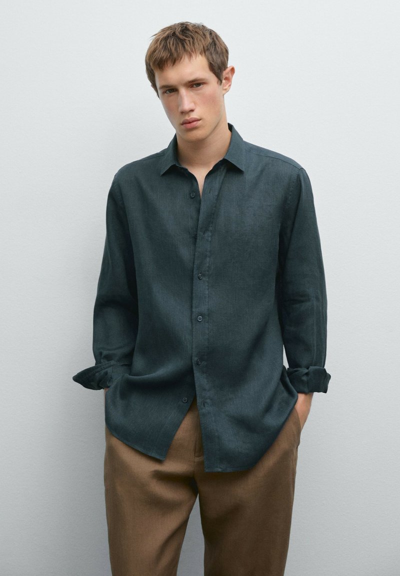 Dark green long-sleeve shirt made of a lightweight fabric, featuring a button-up front, a classic collar, and rolled cuffs. Paired with light brown trousers.