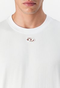 Tricou alb din bumbac, cu decolteu rotund, având o mică decupare ovală și un logo brodat în centrul pieptului.