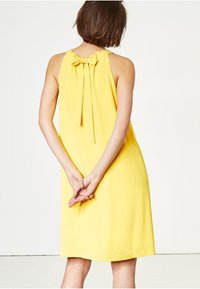 Abito giallo senza maniche con un dettaglio a fiocco legato sulla schiena, caratterizzato da una vestibilità ampia e una trama di tessuto liscia, che arriva sopra il ginocchio.