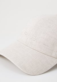 Lys beige caps laget av teksturert stoff med en buet skjerm. Har brodert "BOSS"-logo på frontpanelet.