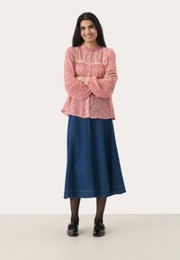Blusa rossa a fiori con dettagli plissettati, abbinata a una lunga gonna in denim. L'outfit presenta una vestibilità comoda e scarpe nere.
