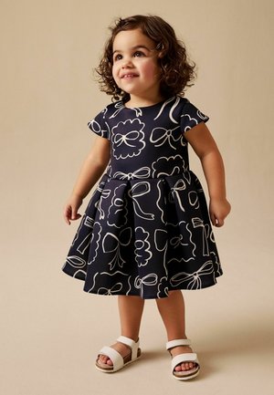 Lächendes Kleinkind mit lockigem Haar, das ein marineblaues Kleid mit weißen Schleifen und weiße Sandalen trägt, steht vor einem beigen Hintergrund.